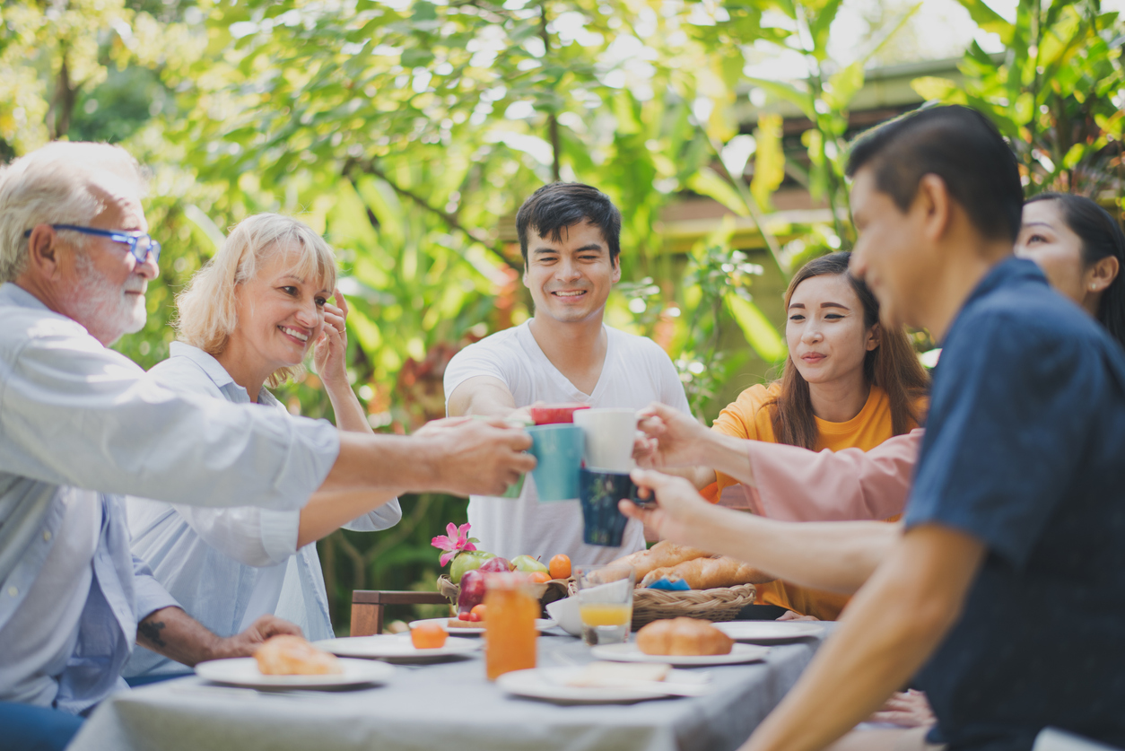 Happy big family have a lunch at outdoor in green garden. Lunch or tea time on picnic table in summer. All people have a clink glasses. Big family outdoor lunch concept.